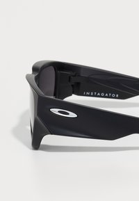 Gafas de sol de plástico negro con lentes grises, que presentan un diseño elegante, acentos laterales distintivos y el nombre de la marca "INSTIGATOR" impreso.