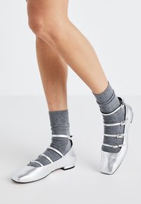 Des chaussures plates argentées avec un bout carré, mettant en avant plusieurs fines brides et un petit talon, assorties avec des chaussettes à cheville grises. Matériau lisse et brillant.
