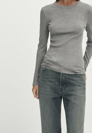 Femme portant un haut gris près du corps à manches longues côtelé, rentré dans un jean noir délavé taille haute, sur un fond uni.