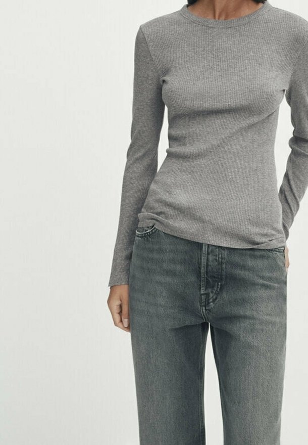 Femme portant un haut gris près du corps à manches longues côtelé, rentré dans un jean noir délavé taille haute, sur un fond uni.