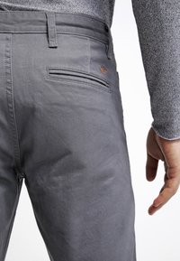 Graue Hosen mit glatter Textur, ausgestattet mit einer Gesäßtasche und einem dezenten, in Kontrastfarbe gestickten Logo. Nahaufnahme des Bunds.