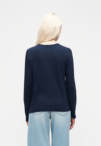 Ξανθό άτομο με την πλάτη στραμμένη, φοράει μπλε ναυτικό μπλουζάκι μακρυμάνικο και ανοιχτόχρωμα τζιν φαρδιάς γραμμής, σε απλό λευκό φόντο.