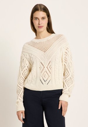 Frau mit glatten braunen Haaren, die einen cremefarbenen Strickpullover mit Rautenmuster und schwarze Hosen trägt, steht vor einem neutralen Hintergrund.