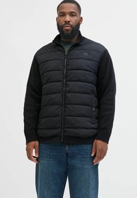 Schwarze Zip-Up-Jacke mit gepolstertem Vorderteil und gestrickten Ärmeln, mit hohem Kragen und dezentem Logoschriftzug auf der Brust.