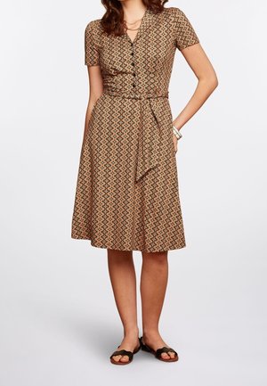Femme portant une robe marron à manches courtes, longueur genou, avec un motif géométrique ondulé, boutonnée devant, ceinture à la taille et sandales noires ouvertes à l'avant.