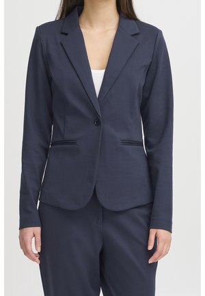 Femme portant un blazer bleu marine foncé à un bouton par-dessus un haut blanc, associé à un pantalon bleu marine assorti, sur un fond clair uni.