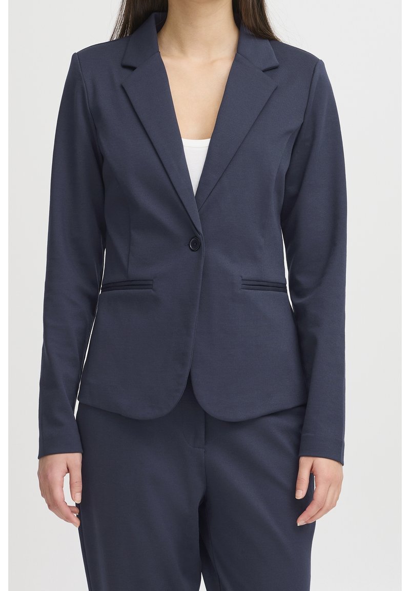 Femme portant un blazer bleu marine foncé à un bouton par-dessus un haut blanc, associé à un pantalon bleu marine assorti, sur un fond clair uni.