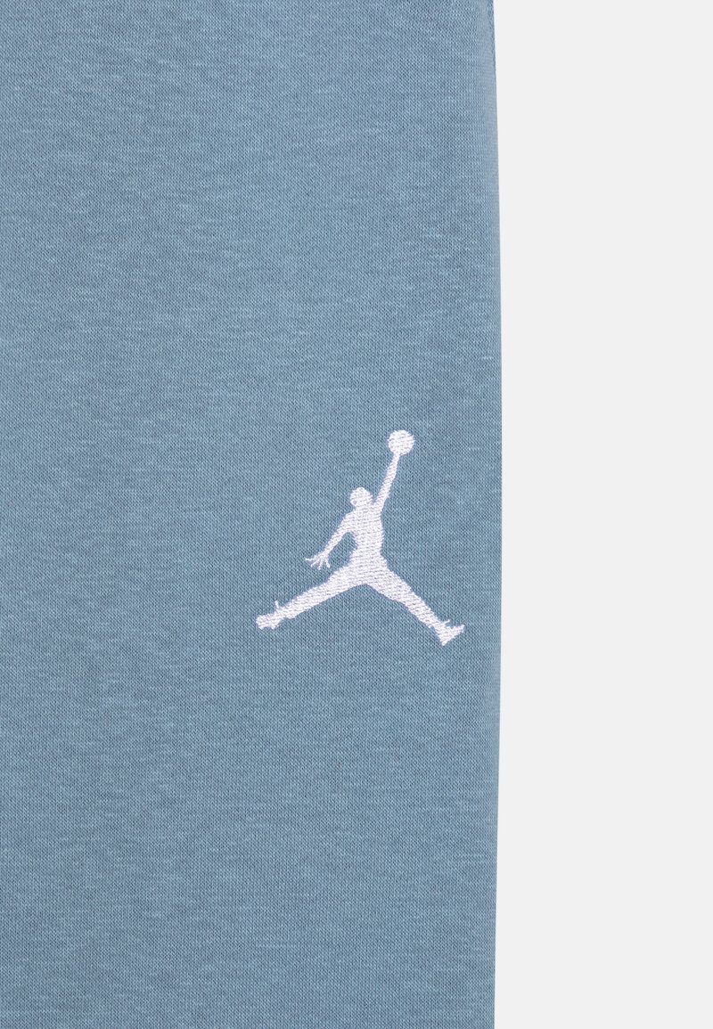 Jasnoniebieska bawełniana tkanina z białym haftowanym logo Jumpman, przedstawiającym postać w powietrzu, o teksturowanym wykończeniu.