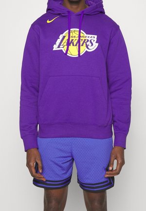 Osoba ubrana w fioletową bluzę z kapturem Los Angeles Lakers z żółtym logo i niebieskie spodenki do koszykówki z czarnymi paskami, stoi na jednolitym tle.