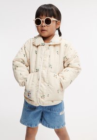 Ljust beige kviltad jacka med blommönster, tryckknappar och två framfickor, tillsammans med denimshorts och runda solglasögon.