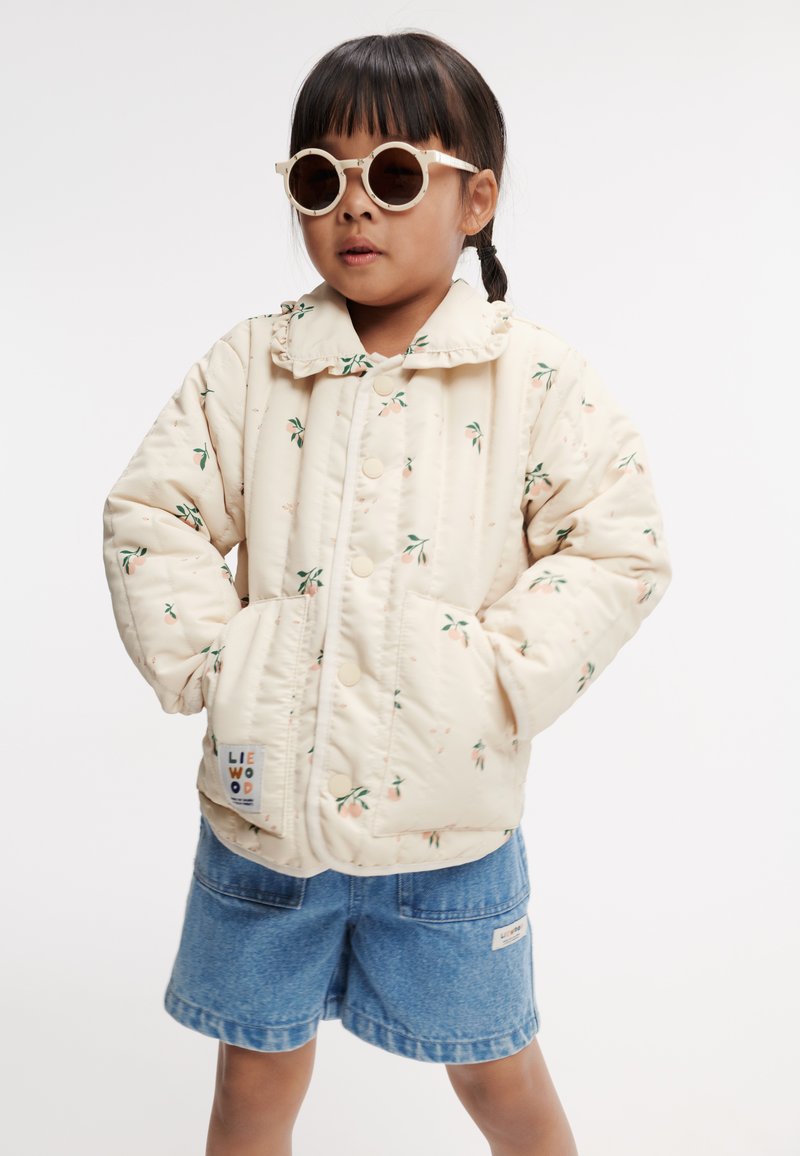 Ljust beige kviltad jacka med blommönster, tryckknappar och två framfickor, tillsammans med denimshorts och runda solglasögon.