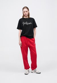 Maglietta nera in cotone con lettering bianco "Jordan", abbinata a pantaloni della tuta rossi. Sneakers con parte superiore grigio chiaro e suola nera.