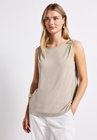 Top sin mangas de color beige claro con un corte relajado, que presenta un herraje decorativo en las tirantes y una textura suave y lisa.