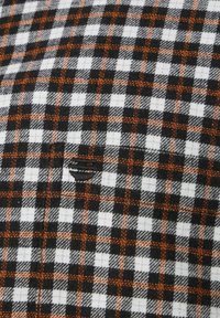 Schwarz-weiße und orange karierte Stoff mit strukturierter Oberfläche. Verfügt über eine kleine Tasche und einen leichten Fransenrand.