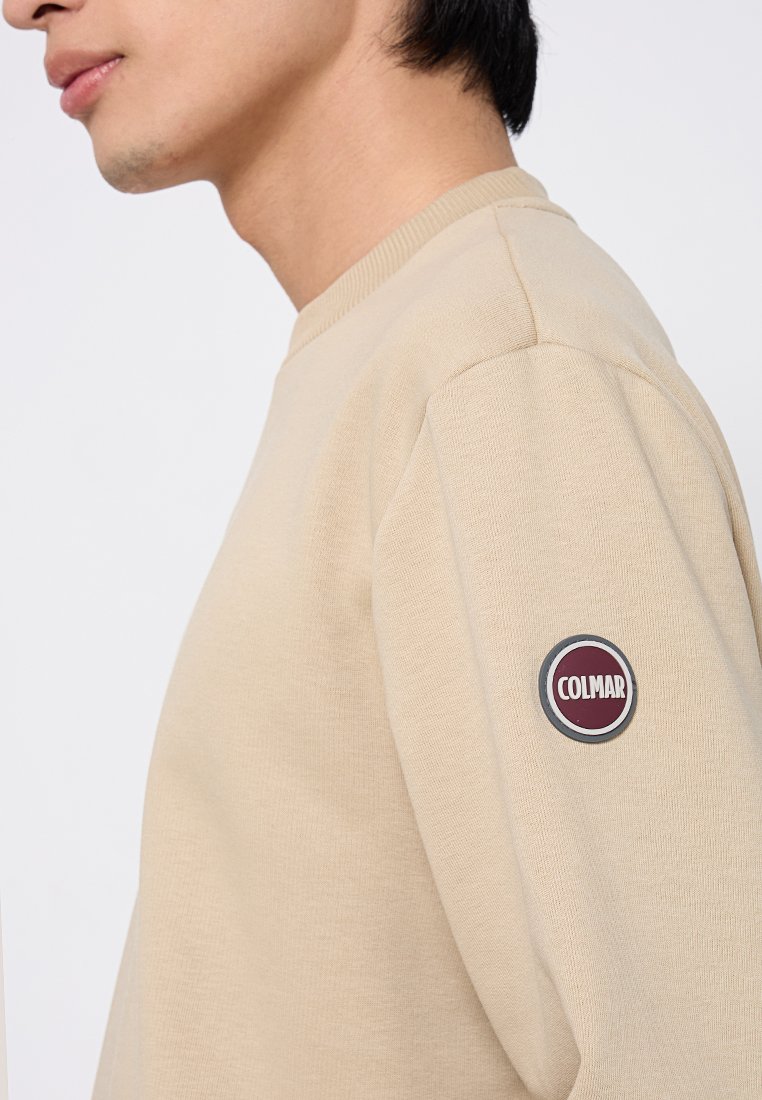Sweatshirt beige con un logo ricamato tondeggiante sulla manica sinistra. Presenta un collo rotondo e polsini a coste. Texture morbida e design informale.