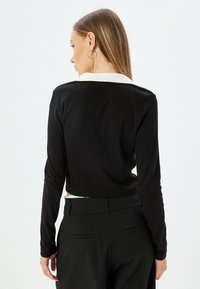 Musta pitkähihaine paita, jossa on kontrastina valkoinen kaulus, valmistettu sileästä kankaasta ja jossa on lyhennetty malli. Yhdistetty istuviin mustiin housuihin.