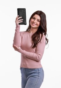 Black leather wallet with perforated design, featuring a zipper. Model holds it in one hand. Ribbed light pink sweater and blue jeans visible.