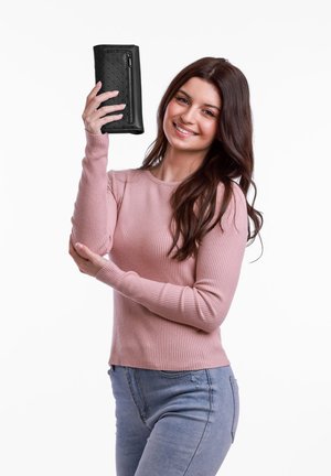 Black leather wallet with perforated design, featuring a zipper. Model holds it in one hand. Ribbed light pink sweater and blue jeans visible.