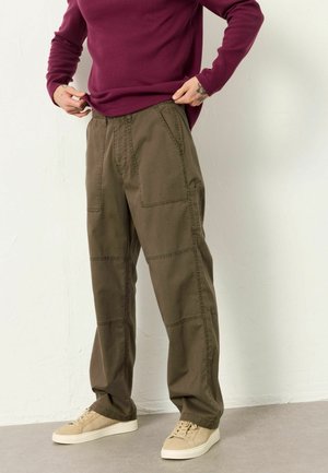 Person wearing olive green cargo pants, beige sneakers, and a long-sleeve dark red shirt, standing against a plain light wall.
