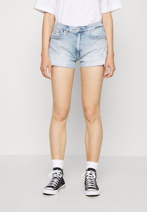Osoba ubrana w jasnoniebieskie szorty dżinsowe, białe skarpetki, czarne sneakersy za kostkę oraz białą oversize koszulkę, stojąca na jednolitym tle.