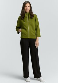 Veste zippée vert olive avec un col montant et un cordon de serrage, associée à un pantalon noir à coupe large et des baskets blanches. Texture de tissu lisse.
