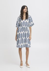 Witte jurk met blauw bloemenpatroon, korte mouwen en elastische taille. Vierkante halslijn, knielengte en lichtgewicht stof.