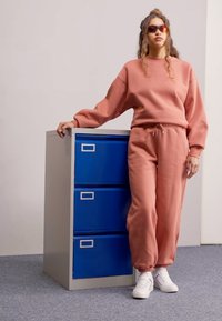 Roze sweatshirt en bijpassende joggers gemaakt van zachte stof, aangevuld met witte sneakers. Blauwe archiefkast op de achtergrond. Zonnebril gedragen.