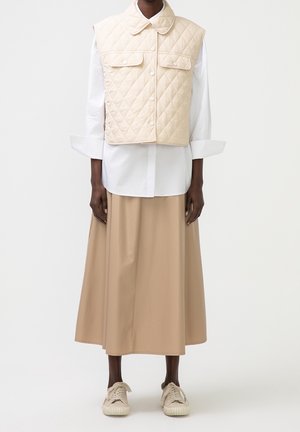 Personne portant un gilet matelassé beige sur une chemise blanche à manches longues, associée à une jupe midi beige et des baskets beiges, debout devant un fond uni.