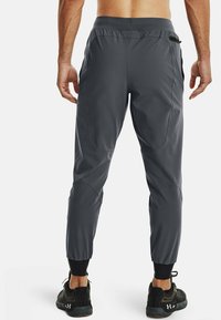 Muž v tmavě šedých sportovních teplácích a černých teniskách Under Armour stojí zády k divákovi na bílém pozadí.