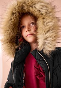 Černá vatovaná bunda s velkou načechranou kapucí s béžovým kožichem. Vnitřní podšívka má barevné vzory. Částečně je vidět bordó vršek.