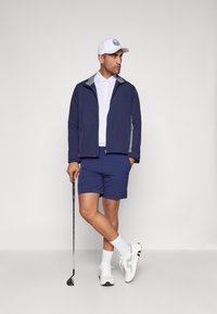 Marinejakke med grå detaljer, hvit polo t-skjorte, blå shorts og hvite sko. Holder en golfklubbe, står mot en nøytral bakgrunn.
