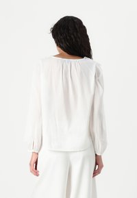 Blusa bianca a maniche lunghe in tessuto leggero, con scollatura arricciata e polsini elastici, indossata con pantaloni bianchi coordinati.