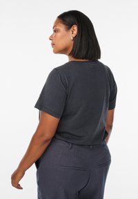 Kvinde med strittende sort hår, iført sølv hoop øreringe, en mørkegrå T-shirt og mørkegrå bukser, som står sidelæns mod en hvid baggrund.