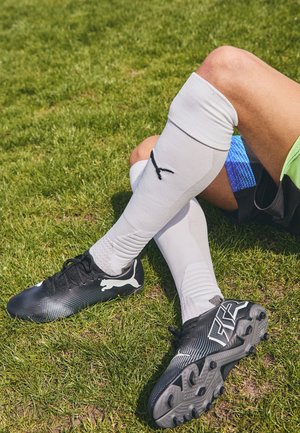 Beine einer Person, die auf Gras sitzt und weiße knielange Socken sowie schwarze Puma Fußballschuhe mit Stollen an den Sohlen trägt.