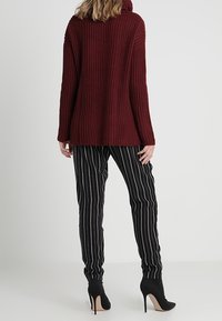 Bordowy sweter z prążkowanej tkaniny z kapturem, zestawiony z czarnymi spodniami w paski. Czarne buty na obcasie. Skupienie na fakturze i kontrastujących wzorach.