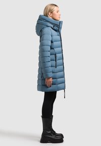 Taivaansininen toppatakki, jossa on korkea kaulus, pystysuuntainen tikattu kuvio, etuvetoketjut ja sivutaskut, yhdistettynä mustiin legginsseihin ja saappaisiin.