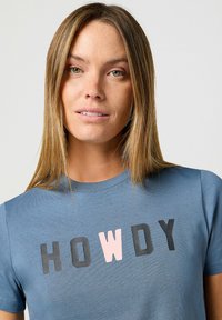 Sininen puuvillainen T-paita lyhyillä hihoilla, jossa on teksti "HOWDY" mustilla ja vaaleanpunaisilla kirjaimilla rinnan kohdalla.