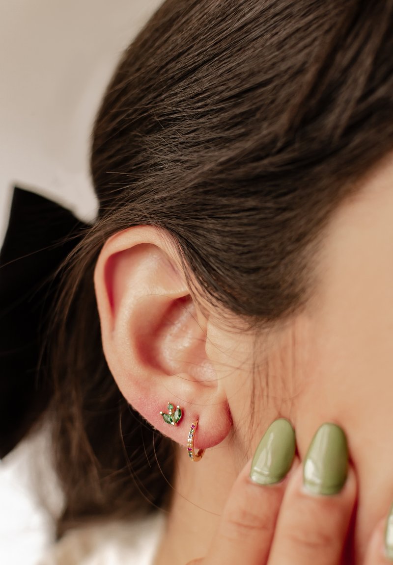 Pendiente de aro dorado con piedras de colores y un pequeño cristal verde, exhibido en la oreja, resaltando detalles y texturas.