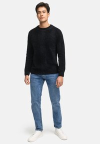 Uomo che indossa un pullover nero con texture, jeans blu e sneakers bianche, in piedi contro uno sfondo bianco uniforme.