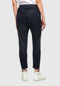 Joggers de denim color azul oscuro con forma ajustada, que cuentan con dos bolsillos traseros y una cinturilla texturizada. Combinados con zapatillas altas blancas.