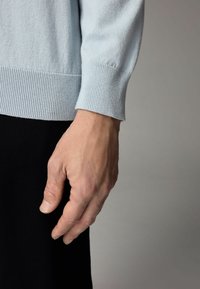 Světle modrý pletený svetr nošený na ruce, s žebrovanými manžetami. Ruka je mírně uvolněná, s viditelnou texturou pleti a nehty.
