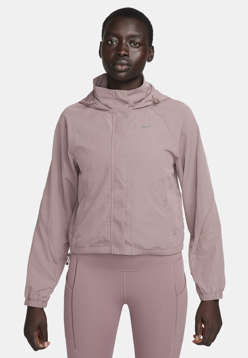 Vrouw in mauve jack met rits en bijpassende leggings, staande tegen een effen lichtgrijze achtergrond, recht vooruit kijkend.