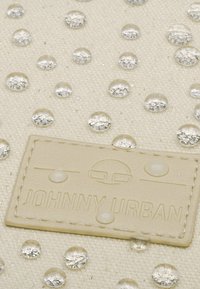 Krém színű textil táska, tiszta vízcseppekkel és egy texturált bézs címkével, amelyen a "JOHNNY URBAN" felirat látható. A körök mérete és elhelyezkedése változó.