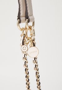 Beige og mørke læder skulderrem med guldfarvet hardware, der har indviklede kæder og cirkulære mærker indgraveret med "G" og "GUESS."