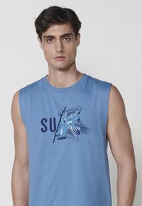 Modra brezrokavna bombažna majica z grafiko "SURF" v odtenkih modre in mornarske barve, okrašena s palmovimi listi ter sproščenim krojem.