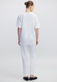 Top blanco de manga corta y pantalones blancos ajustados. Tejido suave con un corte relajado. Zapatos planos negros con acentos decorativos. Fondo neutro.