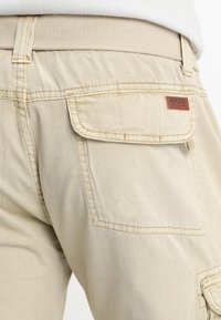 Pantalons cargo en coton beige avec une poche arrière à rabat et une étiquette en cuir marron. La taille est élastique pour plus de confort.