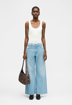 Femeie purtând un top alb fără mâneci și blugi largi albastru deschis, ținând o geantă maro și cizme negre de gleznă, pe un fundal alb simplu.