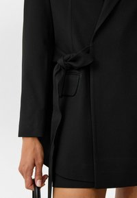 Zwarte wikkeljas met lange mouwen, een strikceintuur en een patchzak. De stof lijkt gestructureerd, wat de strakke vormgeving benadrukt.