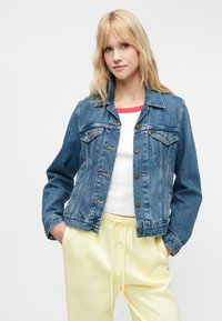 Zilas džinsu jaka ar pogu aizdari, divām krūšu kabatām un sānu kabatām. Nēsāta pāri baltam topam un dzeltenām sporta biksēm, kam ir brīva piegriezuma stils.